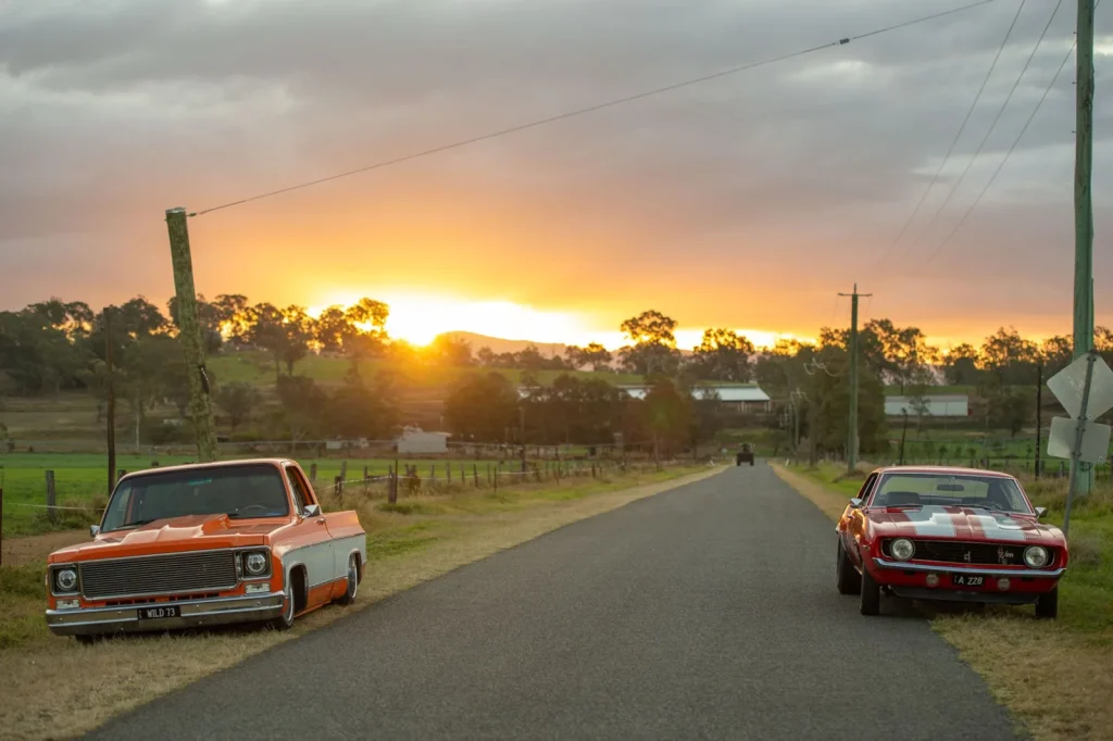 Lowered orange pickup and red muscle car parked either side of a country road at sunset